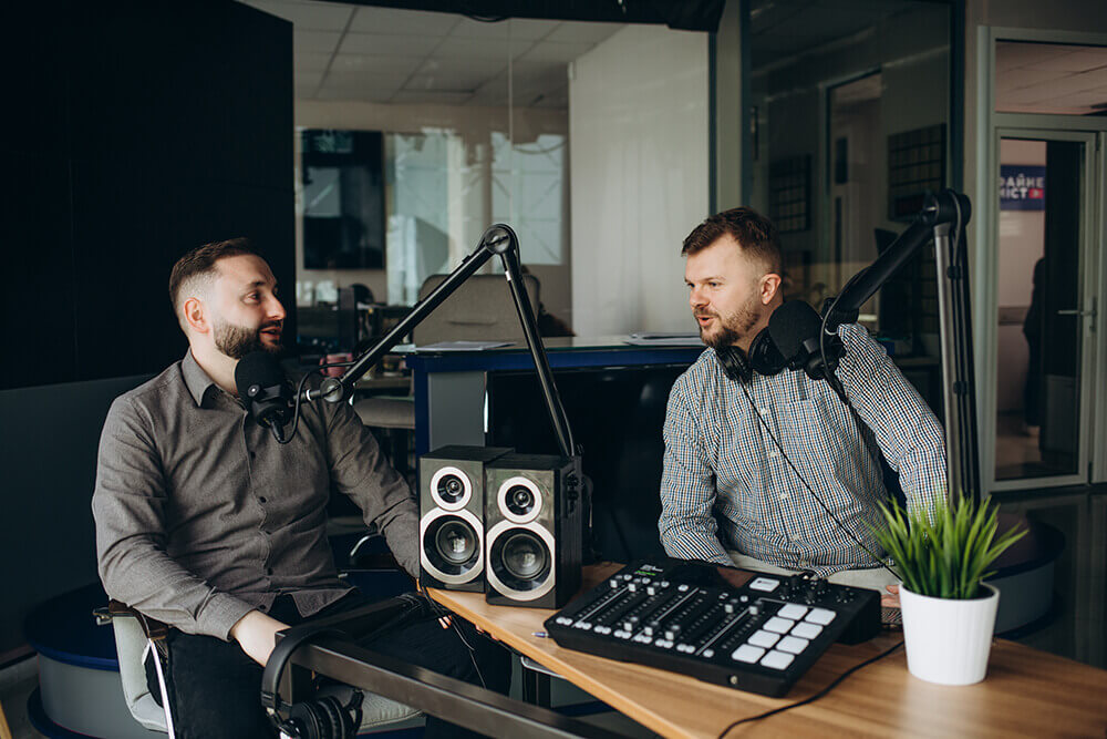 Two smiling radio hosts in a broadcasting studio, engaged in conversation while recording a podcast.